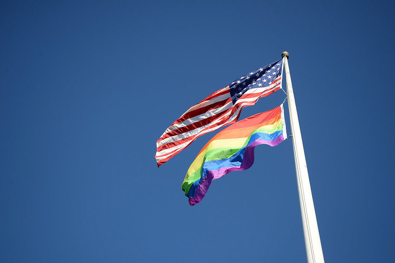 Pride Flag and USA Flag flying from flagpole against blue sky